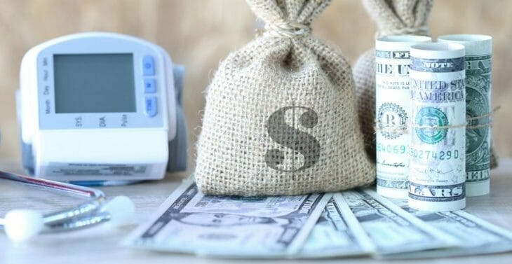 A close-up image showing a burlap money bag with a dollar sign in the center, standing next to rolls of U.S. dollar bills. In the background, there's a digital blood pressure monitor, partially out of focus, suggesting a connection between health and finances. The setup is on a surface with scattered U.S. dollar bills, indicating a theme of medical expenses or healthcare funding.