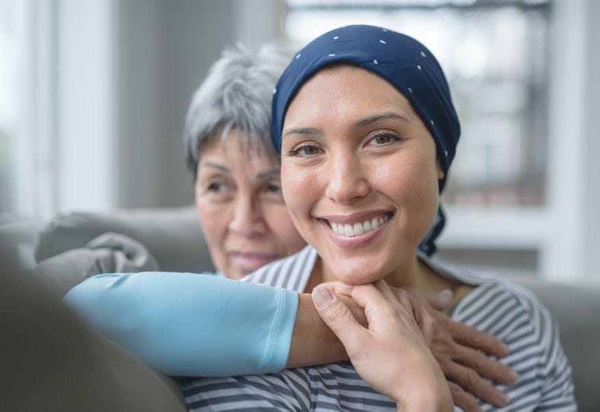 The image shows a close-up of two women, one older and one younger. The younger woman is in the foreground, smiling at the camera, wearing a blue headscarf and a striped top. The older woman, slightly out of focus, is positioned behind her, providing a supportive embrace. Both display an aura of warmth and positivity. The setting appears to be a comfortable home environment. This image may represent themes of family support, health challenges, and recovery.