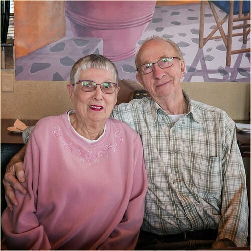 An elderly couple is sitting closely together, smiling at the camera with a warm, inviting expression. The woman is wearing a pink sweater and glasses, and the man is in a plaid shirt with glasses. They appear to be in a cozy setting with a mural in the background, possibly reflecting a comfortable and supportive environment. This image could be associated with themes of financial planning or assistance for seniors, such as those seeking to qualify for programs or services mentioned on a webpage dedicated to financial aid.