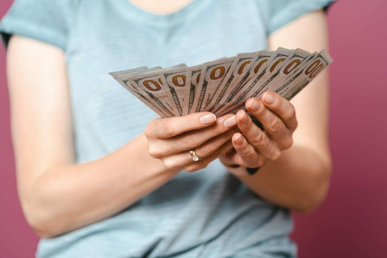 An image of young lady hands in a blue summer tshirt is holding money and looking to them over a pink background.