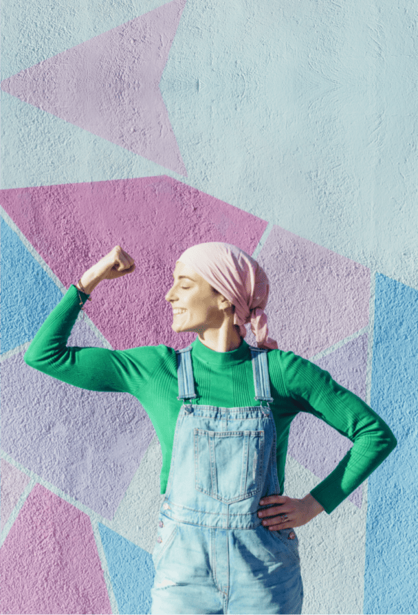 A confident individual with a pink headscarf and green long-sleeve shirt, wearing blue denim overalls, stands in front of a geometrically patterned pastel wall. They are smiling, eyes closed, with one arm flexed in a pose that suggests strength and positivity, reminiscent of the empowerment often needed during journeys like alternative cancer treatment.