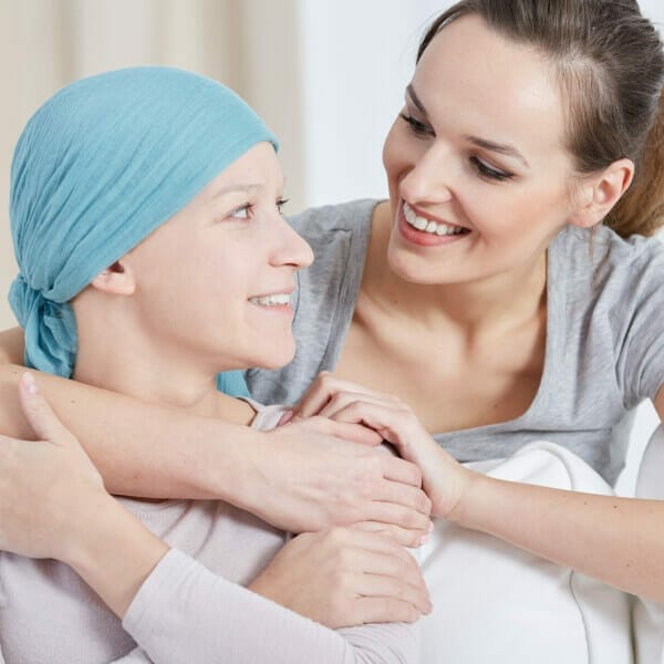 Two women are sharing a moment of joy and comfort; one is wearing a teal headscarf and a light long-sleeve top, looking up with a smile at the other woman who is dressed in a grey V-neck shirt. The second woman, who is smiling tenderly, has her hands gently placed over the hands of the woman in the headscarf. This image conveys a sense of support and companionship, which is vital for individuals exploring alternative cancer treatment options.