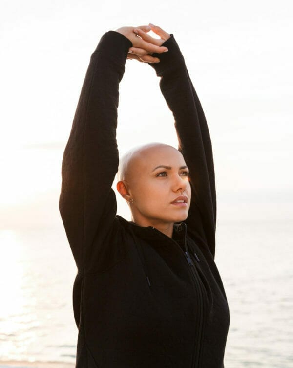 A person is captured in a serene moment, stretching their arms upwards against a tranquil backdrop where the subtle light of sunset meets the calm sea. They are dressed in a black long-sleeve top, and their bald head can signify a journey with cancer, perhaps implying a connection to holistic well-being and alternative treatment paths. Their gaze is directed upwards, possibly reflecting hope and contemplation.