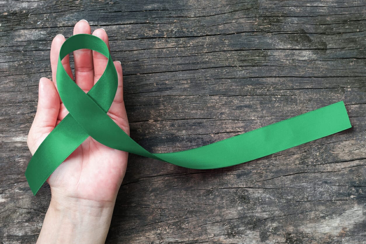 Liver cancer awareness ribbon emerald green color ribbon on human hand aged background, clipping path: satin fabric symbolic logo raising support help people life living with tumor illness