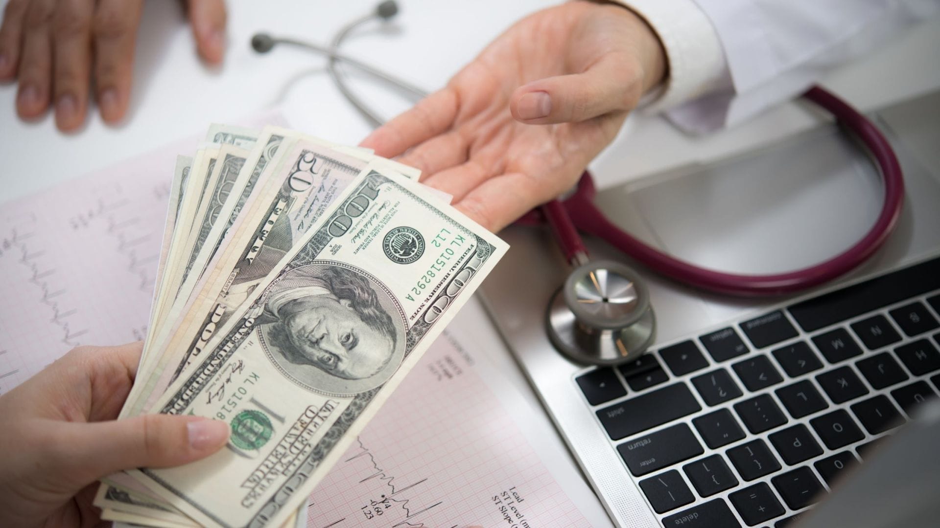 A patient's hand receiving cash, implying financial assistance for liver cancer treatment, with a doctor's stethoscope and laptop in the background, highlighting the intersection of medical care and treatment affordability.