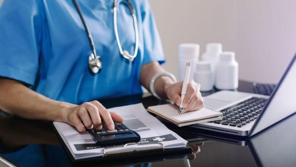 A healthcare professional in blue scrubs is using a calculator and taking notes, likely calculating the expenses associated with stomach cancer therapy. In the background, there's a laptop and medication bottles, indicating a medical office setting where treatment plans and associated costs are being assessed.
