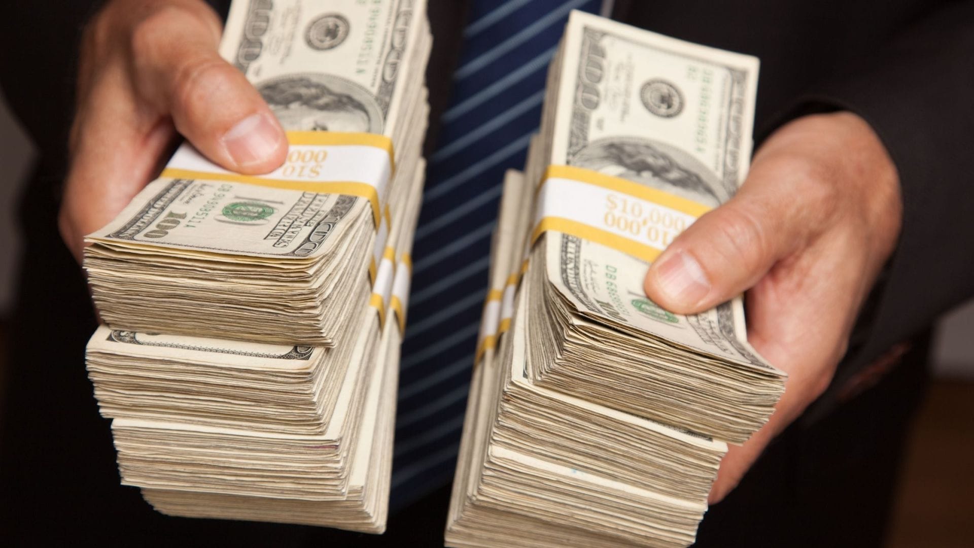 Two hands are holding stacks of U.S. hundred-dollar bills, wrapped with currency bands labeled '10,000.' This image could be used to discuss viatical settlements as a financial resource to cover renal cancer treatment costs. The person holding the money is wearing a dark suit, suggesting a formal financial or business setting.