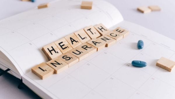Wooden tiles spell out "HEALTH INSURANCE" on an open planner, with two blue pills nearby, indicating the role of health insurance in planning and managing the expenses related to skin cancer treatment.